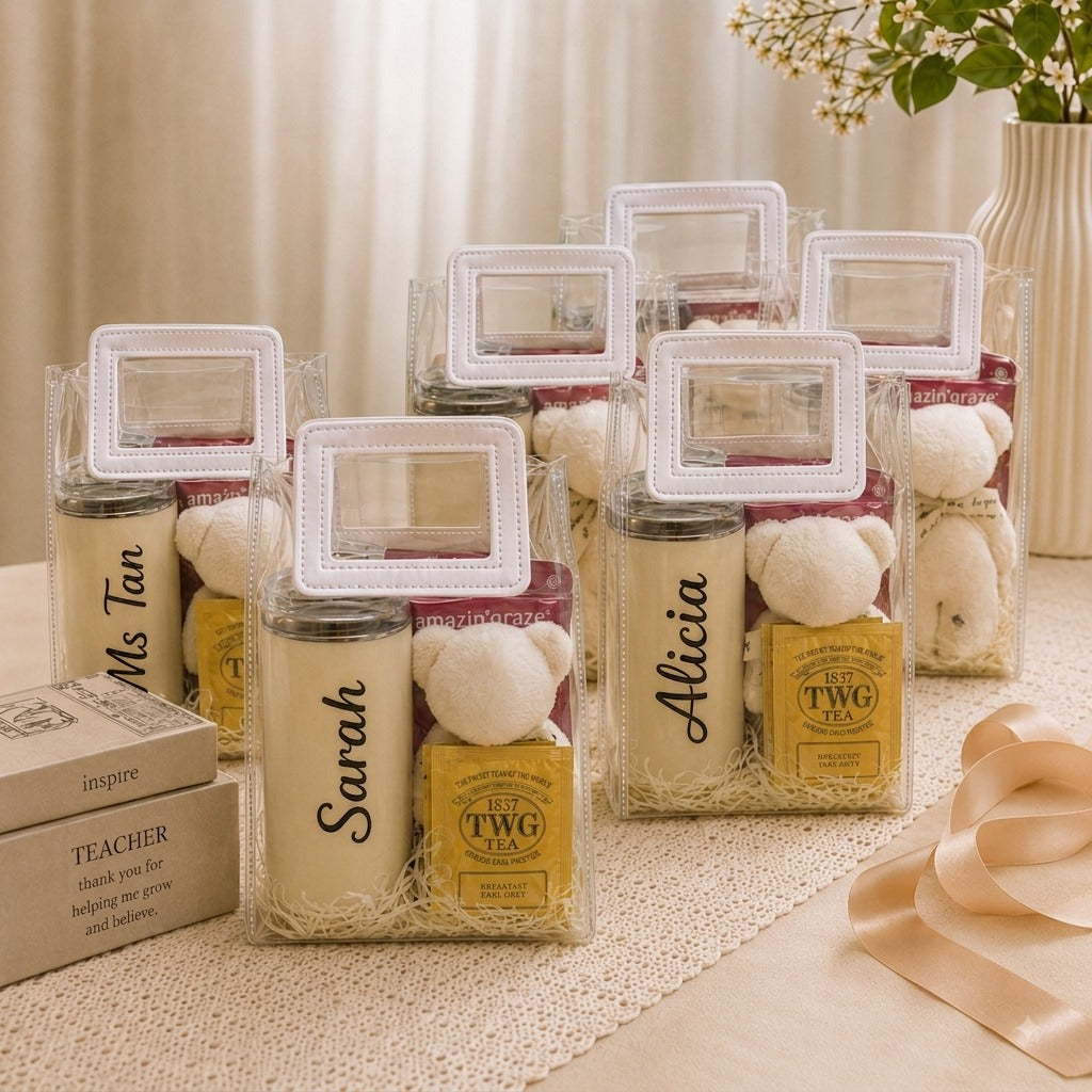 Six personalized TWG tea gift sets in clear plastic gift bags with white handles, arranged on a beige table runner with decorative books and a ribbon. The sets include a cream-colored TWG tea tin, a small white plush bear, and a yellow TWG tea box.