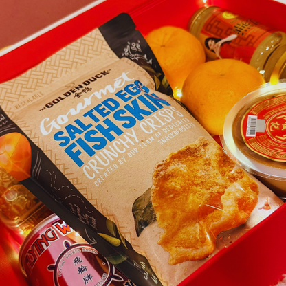 Festive CNY care package contents with mandarin oranges and celebration treats. Packaged food items including a box of Gourmet Salted Egg Fish Skin Crunchy Crisps on a red surface.