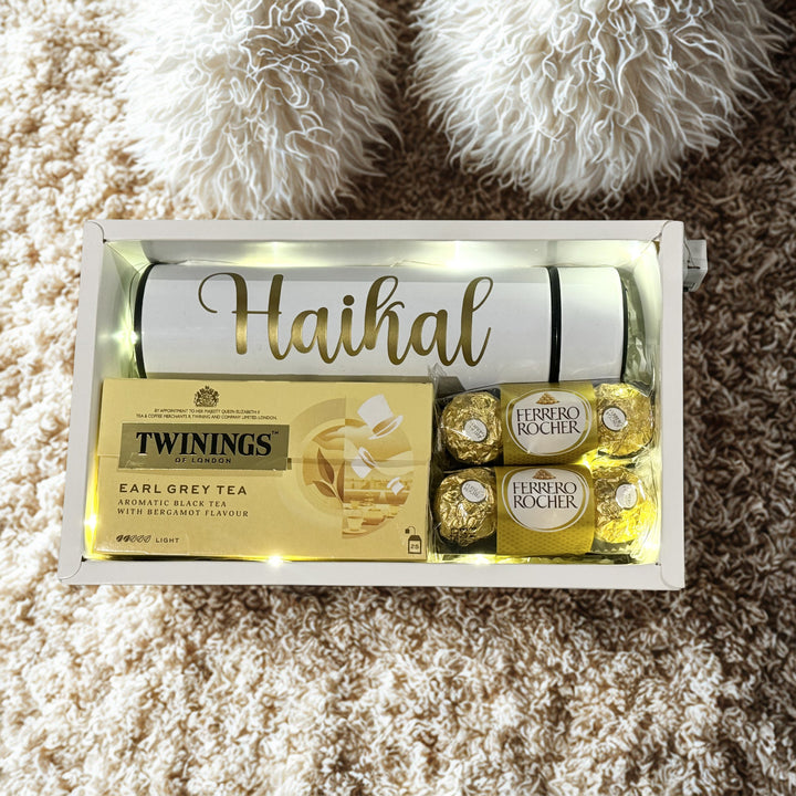 Gift set with Twinings tea, Ferrero Rocher chocolates, and a bottle labeled 'Haikal' on a textured surface.
