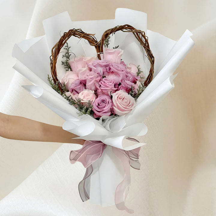 A handcrafted heart-shaped wicker frame bouquet with soft pink roses and greenery, wrapped in white paper with a sheer pink ribbon.