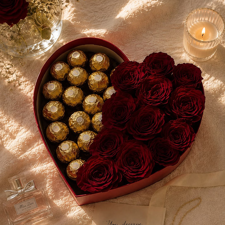 A heart-shaped gift box filled with red roses and Ferrero Rocher chocolates, styled on a textured cream fabric with a lit candle, a small perfume bottle, a card, and a necklace nearby.
