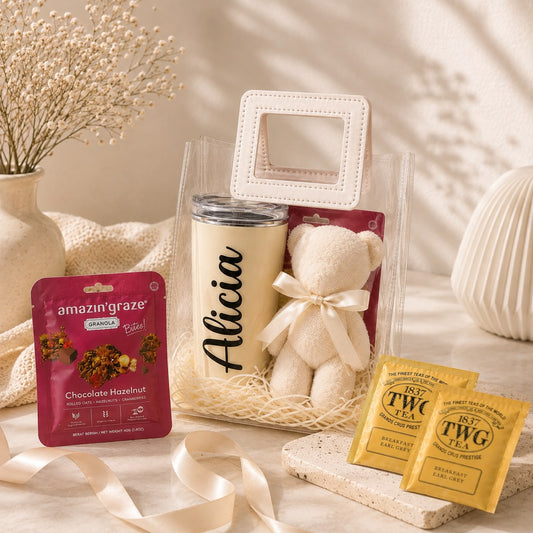 A curated gift set in a clear tote bag with a cream teddy bear, a cream insulated tumbler with the name 'Alicia' in black script, a pink Amazon Prime box, and two tea packets, styled on a neutral fabric surface with dried flowers and soft shadows.